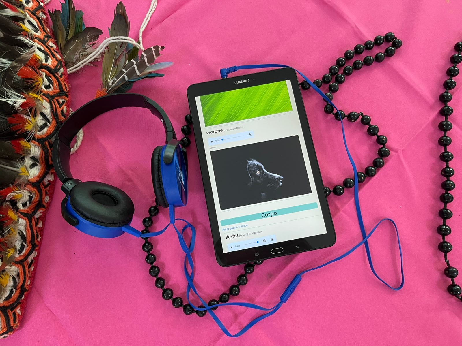Sobre uma toalha rosa vibrante, um tablet Samsung exibe um aplicativo digital com imagens e palavras em idioma indígena. Ao lado, há um par de fones de ouvido azul e um cocar feito com penas naturais, representando a interseção entre tecnologia digital e culturas originárias. Um colar de contas pretas completa a composição, sugerindo a valorização dos saberes ancestrais aliados à inovação.